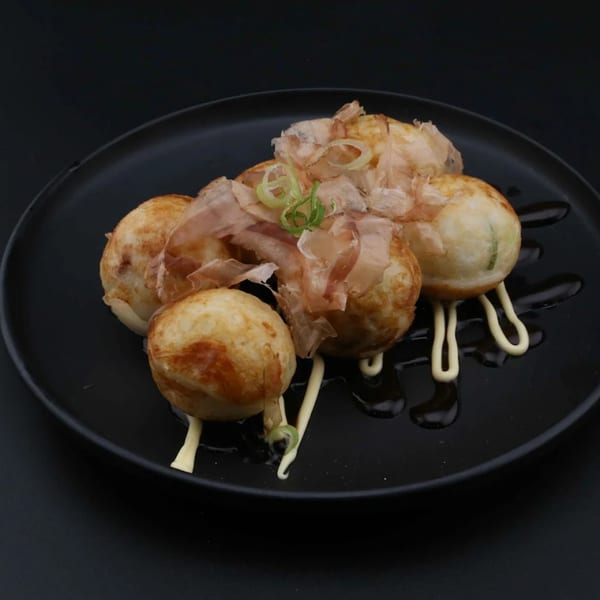 Golden-brown takoyaki balls topped with bonito flakes, green onions, and mayonnaise on a black plate.