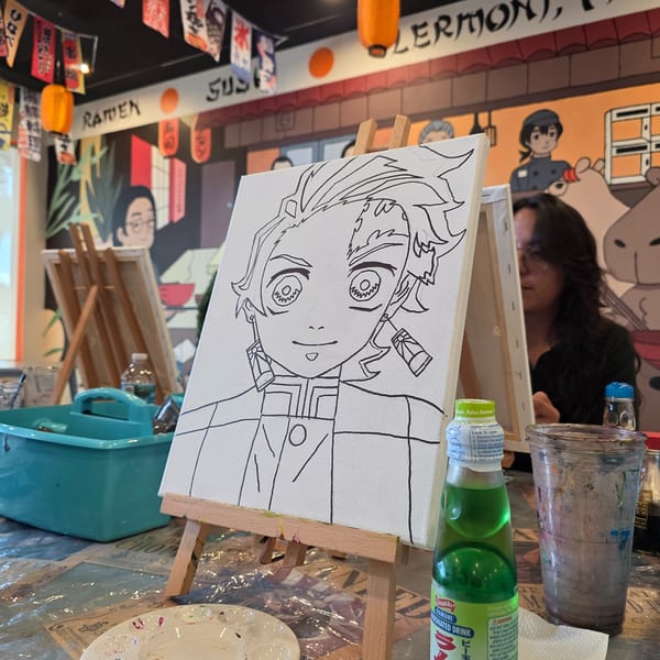 Anime character drawing on an easel with a drink and paint palette, a person painting in the background.