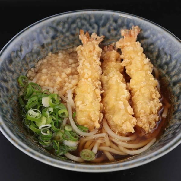 A bowl of udon noodles with three pieces of shrimp tempura and chopped green onions.