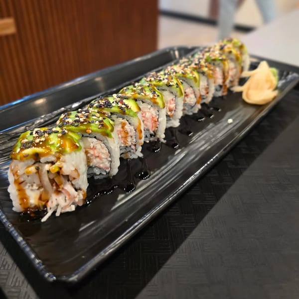A long black plate of sushi rolls topped with avocado, sesame seeds, and sauce, with ginger on the side.