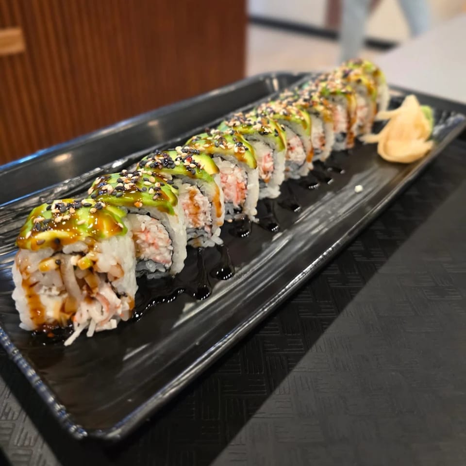 A long black plate of sushi rolls topped with avocado, sesame seeds, and sauce, with ginger on the side.