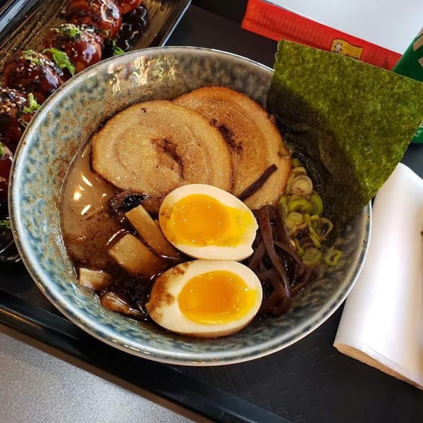 A bowl of ramen with pork, soft-boiled eggs, scallions, and seaweed, next to a plate of takoyaki.